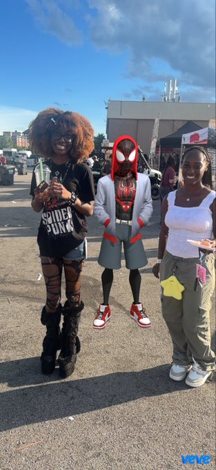 Three people posing at an outdoor event, with the middle person dressed in a Spider-Man costume, labeled 'VOXIC' in the corner.