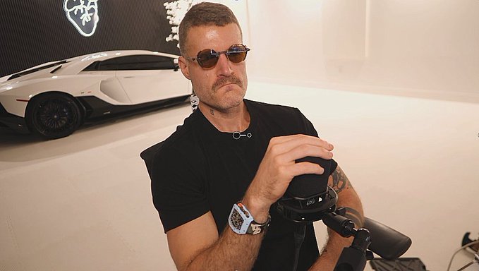 A man with a mustache wearing sunglasses and a black t-shirt, holding a camera in a room with a white Lamborghini Aventador displayed in the background.