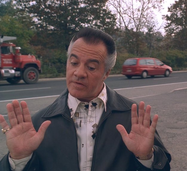 A middle-aged man in a leather jacket holds up both hands with palms facing forward, standing outdoors with a red truck and red minivan visible in the background.