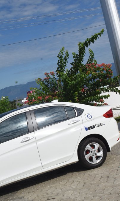 A white hatchback car parked in front of a flowering bush under a blue sky, featuring a blue circular logo and 'base.' text with a bar chart graphic on the rear door.