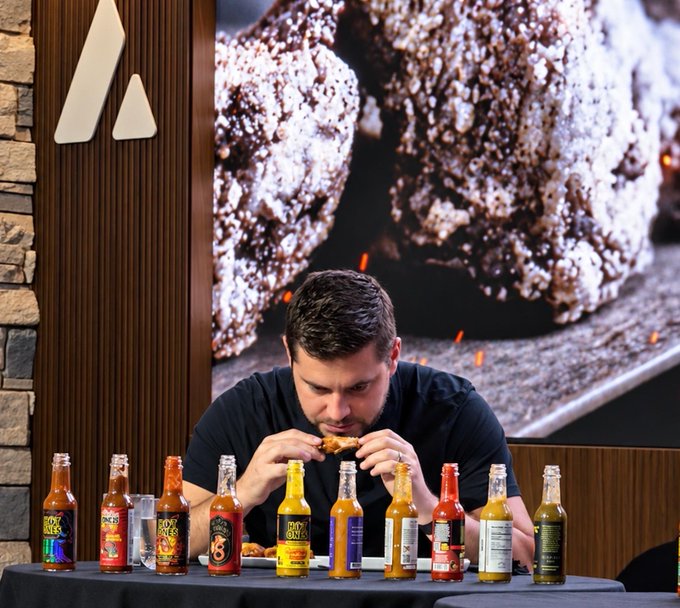 A bearded man in a dark t-shirt bites into a chicken wing at a table lined with a dozen hot sauce bottles labeled "Duke's," "Soy Moses," and others, with a large photo of spiced wings in the background—capturing the wing-eating contest moment before Bo Hines of Tether takes the stage.
