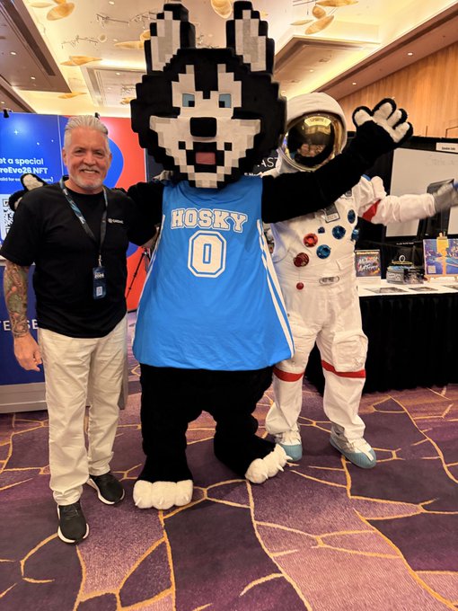 A man in a black Microsoft shirt stands alongside a pixelated Husky mascot wearing a blue "HOSKY 0" jersey and a white astronaut suit at what appears to be a crypto/space-themed event. The image ties into the post by promoting the $HOSKY token through the mascot, while the astronaut's pose ("reaching out") cleverly plays on the "secure your assets" message in the caption.