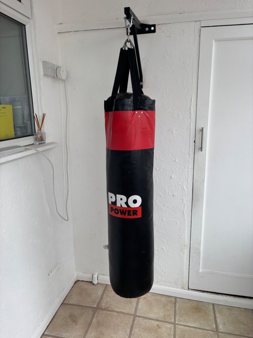 A black and red 'PRO POWER' heavy punching bag hanging from a wall-mounted bracket in a room with white walls and tiled flooring.