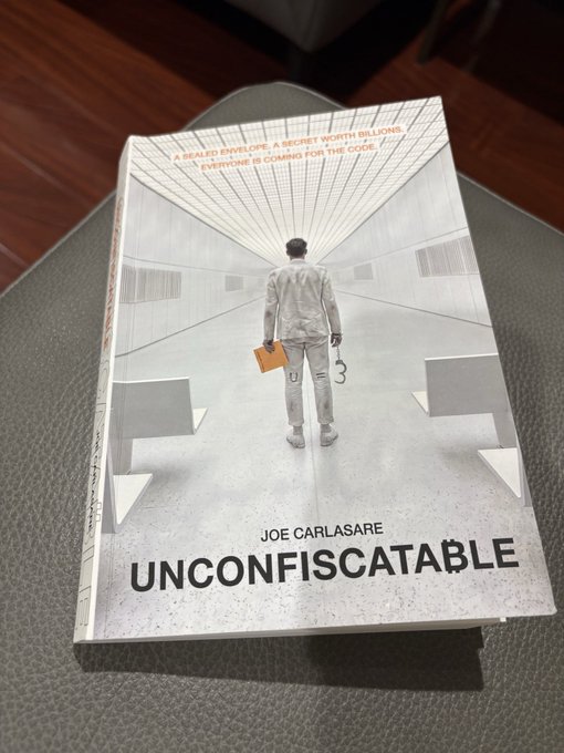 A book titled "Unconfiscatable" by Joe Carlasare rests on a dark surface, featuring cover text: "A SEALED ENVELOPE. A SECRET WORTH BILLIONS. ONE TARGET." and "EVERYONE IS COMING FOR THE CODE." The cover illustration shows a figure in white holding an orange folder and what appears to be a cane.