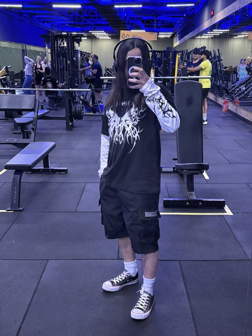 A mirror selfie taken in a gym showing a person wearing a black graphic t-shirt over a white long-sleeve shirt, black cargo shorts, and Converse sneakers, while taking the photo with their phone.