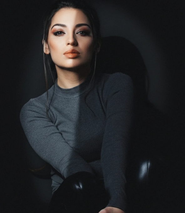 A portrait shows a woman with dark hair wearing a gray long-sleeved top, posed against a dark background.
