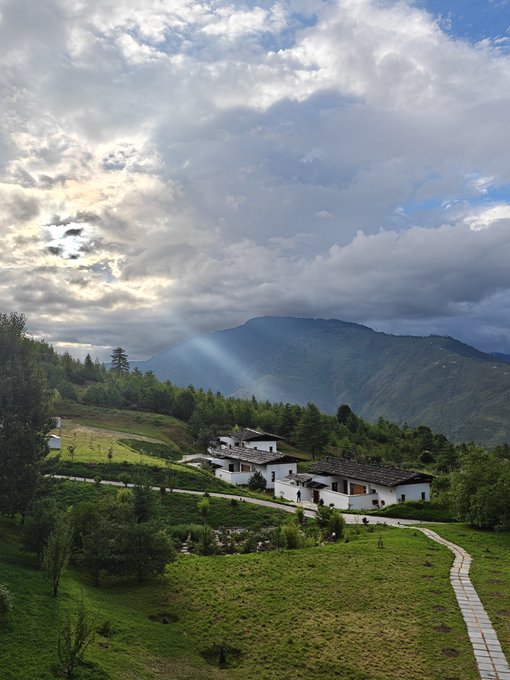 A lone white-walled house with a traditional dark roof sits nestled on a lush green hillside, with mist-draped mountains rising in the background under a dramatic sky where sun rays pierce through the clouds. The image reflects Bhutan's serene and tranquil landscape, which aligns with the post's description of the country as a "magical kingdom" known for its safety, natural beauty, and high national happiness index.