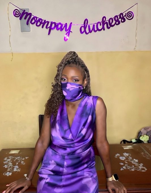 A woman with braided hair wearing a purple camouflage dress and matching face mask sits at a desk displaying jewelry, with a purple 'moonpay duchess' banner on the wall behind her.