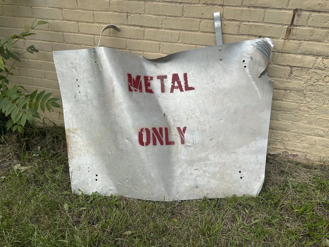 A dented metal panel leans against a brick wall outdoors, stenciled with the red text 'METAL ONLY'.