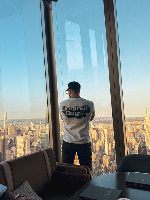 A man wearing a white 'Doginal Dogs' t-shirt and a baseball cap standing in front of floor-to-ceiling windows overlooking the New York City skyline, with an Empire State Industries book on a nearby table.