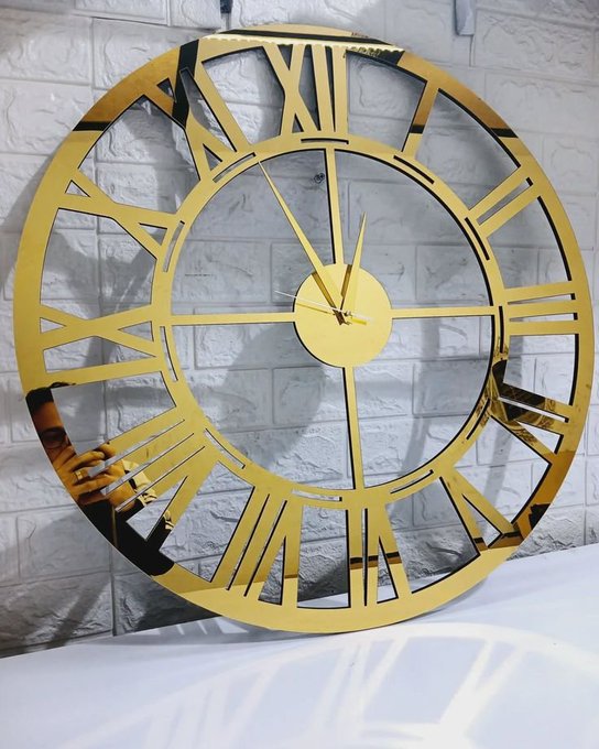 A large decorative gold wall clock with Roman numerals and cut-out hands is displayed against a white brick wall, casting a shadow.