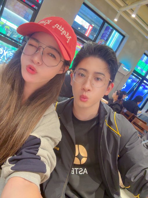 A couple takes a selfie at what appears to be a restaurant or cafe; the woman wears a red 'Mardi Mercredi' cap and oversized glasses, while the man wears a dark jacket and round glasses, both making pouty faces at the camera.