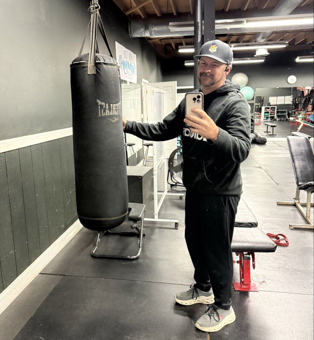 A man in a black hoodie and cap takes a mirror selfie in a gym while holding a hanging black heavy bag; gym equipment and exercise balls are visible behind him.