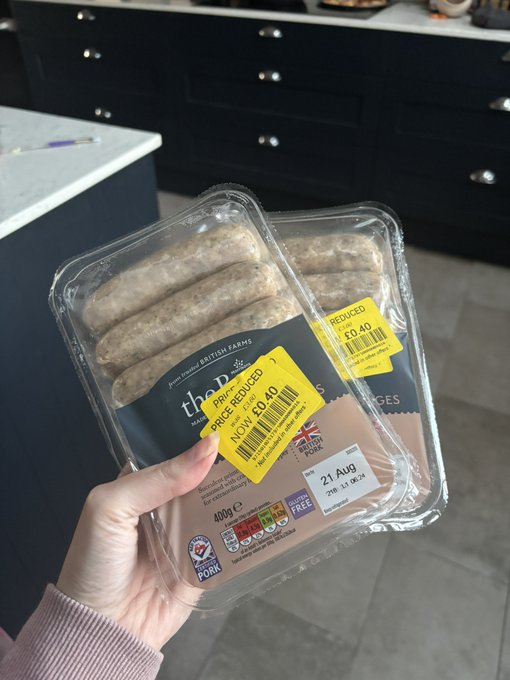 A hand holds two packs of British Farms sausages from a convenience store, both marked down with "PRICE REDUCED" yellow stickers showing £0.40 each, with a sell-by date of 21 Aug — illustrating the post's tip about scoring discounted meat bargains at late-night mini supermarkets to fill a freezer and save money.