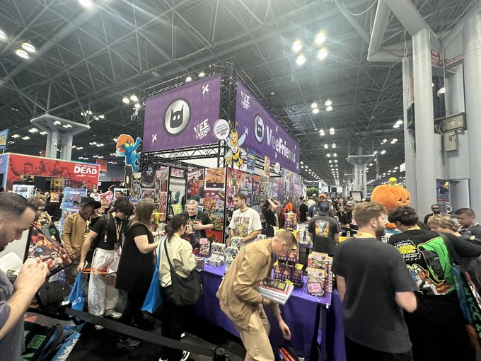 A crowded convention floor featuring a purple-themed booth with "Vee Friends" branding and X logo, where attendees browse merchandise around displays decorated with cartoon characters and Halloween props.