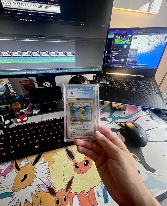 A person holds up a CGC-graded Japanese Pokémon card (a graded 9 Poliwag) in front of a dual-monitor gaming setup with a Dell display, a video editing timeline, and a Pokémon-themed desk mat featuring Eevee evolutions.
