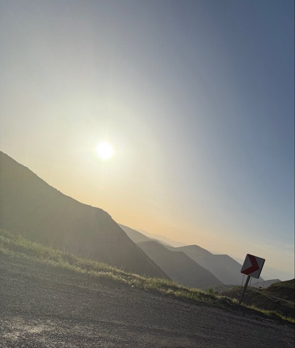 A winding mountain road descends through hazy hills at sunrise, with a right-curve road sign indicating a sharp bend. The scenic yet perilous descent visually captures the nervous anticipation behind the question "Can I GET Gm," symbolizing a cautious ride toward something desired.