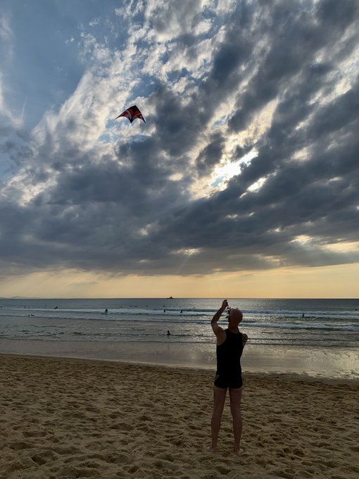 A woman stands on a sandy beach at sunset, flying a kite under dramatic cloudy skies.