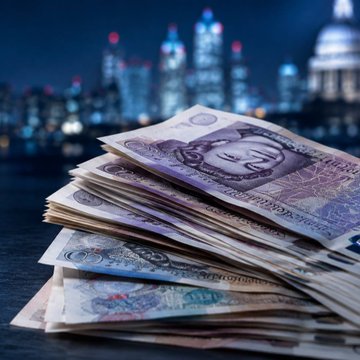 A stack of £50 notes sits on a dark surface with a blurred nighttime city skyline in the background. The image visually reinforces the news of a record 240 UK individuals reporting over £1 million in crypto capital gains for the 2024-2025 tax year.