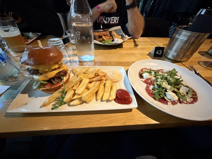 A restaurant table holds a burger with fries and ketchup, a beef carpaccio dish, drinks, and a table number '43'.