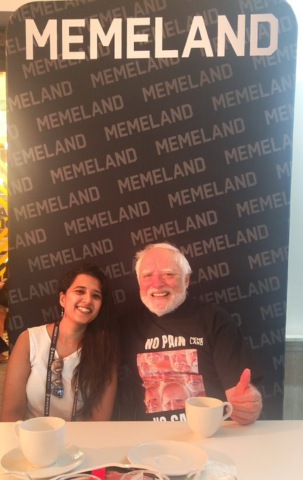 Two people posing in front of a backdrop covered with 'MEMELAND' logos, sitting at a table with coffee cups.