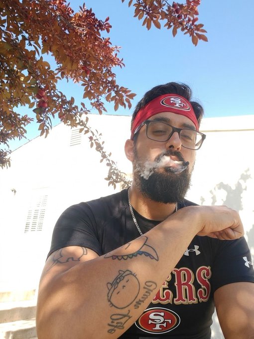 A bearded man wearing a red 49ers headband and black 49ers shirt exhales smoke while flexing his tattooed arm, with red leaves visible overhead.