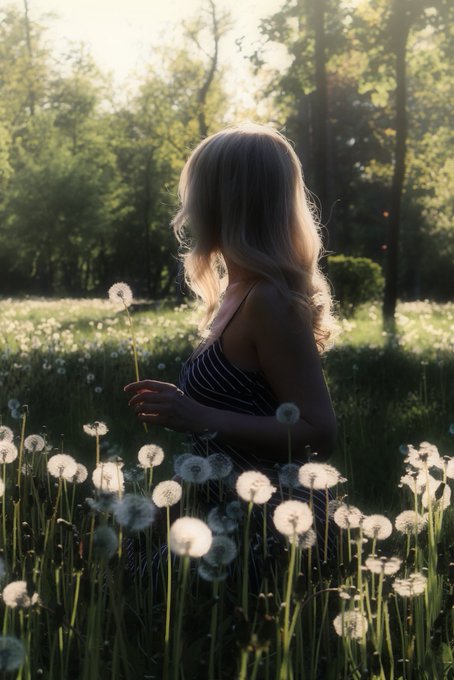 A blonde woman in a striped dress stands in a sunlit field of dandelions, holding one of the fluffy seed heads as sunlight filters through trees in the background.