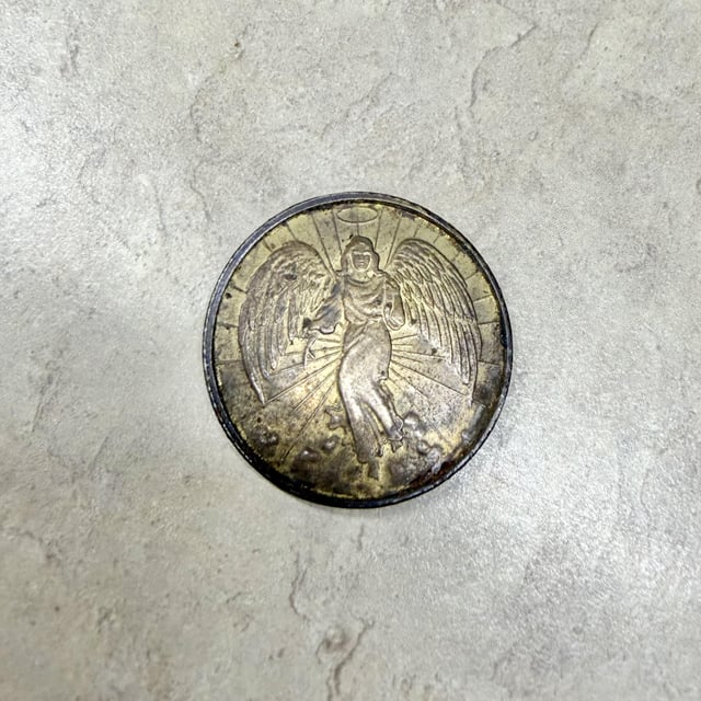 A silver coin or medallion featuring a relief design of an angel with outspread wings stands upright on a textured gray surface.