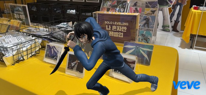 A figurine of an anime character in a blue outfit wielding a knife is displayed on a yellow tablecloth at a Solo Leveling (나 혼자만 레벨업) merchandise stand, with a "veve" logo in the corner.