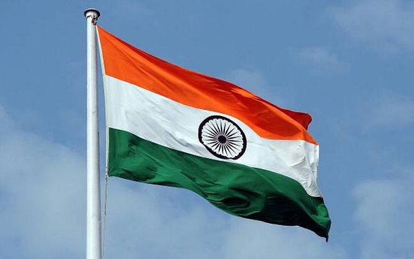 The Indian national flag (tricolor of saffron, white with the Ashoka Chakra, and green) waving on a flagpole against a blue sky.