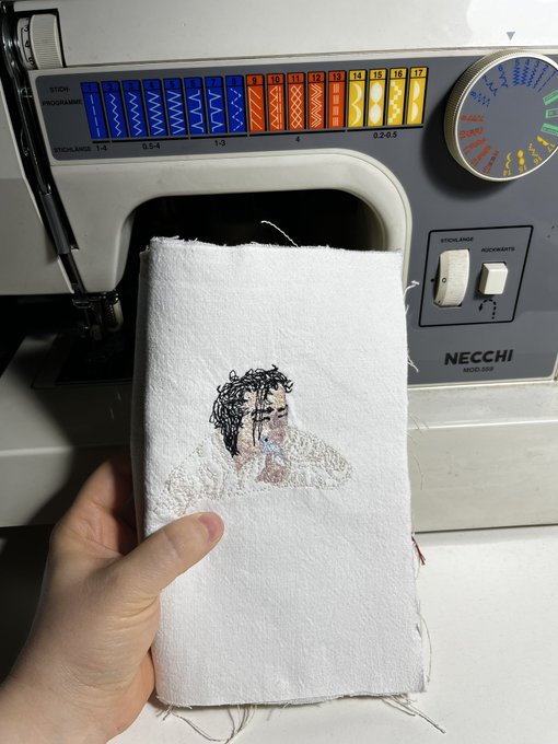A hand holding a small piece of white fabric with a sketch of a person, positioned in front of a Necchi sewing machine.