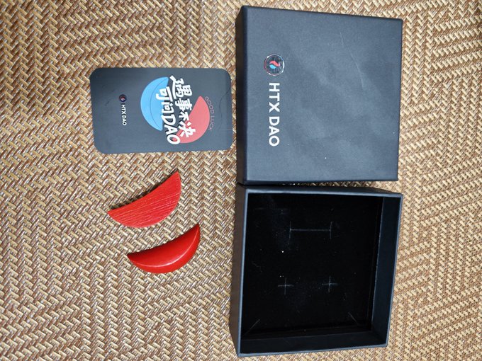 A photo showing an "HTX DAO" branded black gift box, a black card with "HTX DAO" and Chinese text "越织越猛", and two red crescent-shaped items arranged on a woven mat.