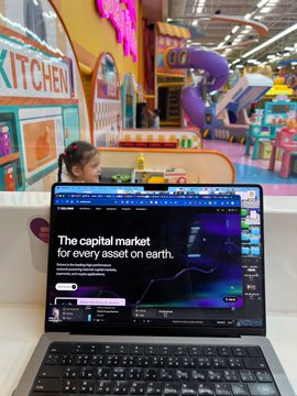 A woman working on a laptop displaying 'The capital market for every asset on earth' in what appears to be a colorful indoor playground or children's play area