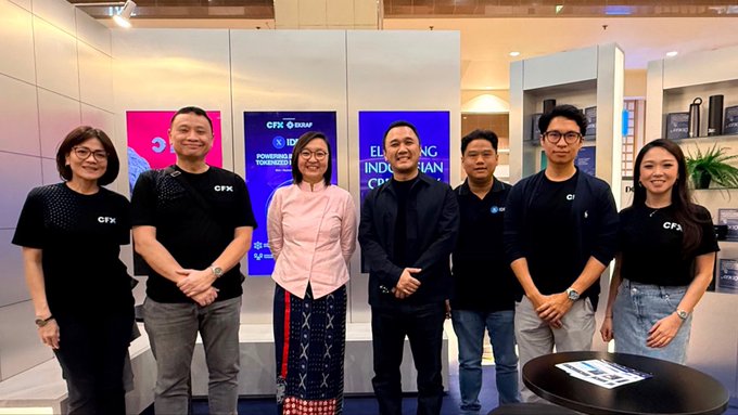 A group of seven people poses at what appears to be a CFX (Corporate Fitness Expo) event, with signage mentioning 'Powering Indonesian CFG' and partners like AXA and ID; one woman wears a traditional pink kebaya.
