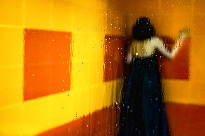 A blurry, impressionistic image of a figure in a dark dress standing in a yellow and orange room, viewed through a rain-streaked or frosted glass surface.