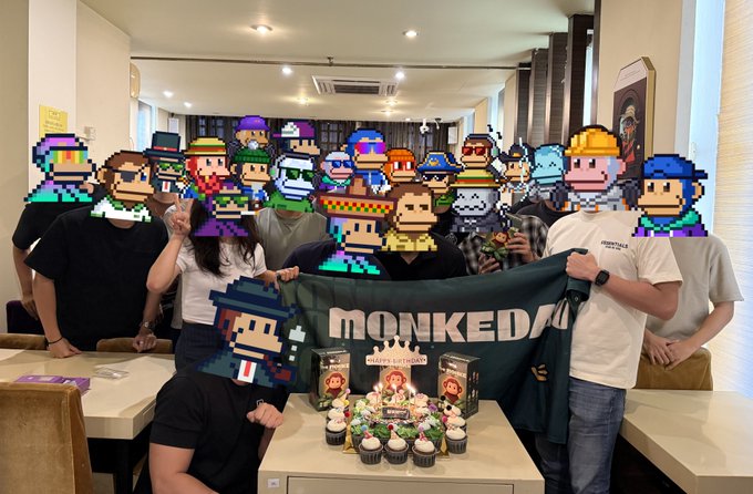 A group photo of people in a cafe with pixel-art face filters, holding a 'MONKEDA' banner above a table with a cake and 'HAPPY BIRTHDAY' decorations.