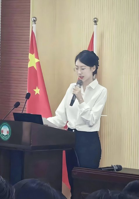 A young woman with glasses and dark hair speaks into a microphone while standing at a podium, with a Chinese flag visible in the background.