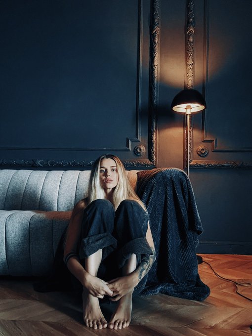 A moody portrait of a young woman with long blonde hair sitting barefoot on a herringbone wood floor in front of a grey couch, with a warm lamp illuminating the dark wall behind her.