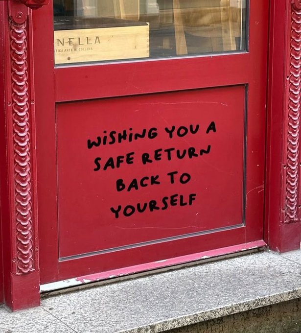 The image features a red door with black text that reads "Wishing you a safe return back to yourself" written on it, which appears to be a message or greeting for someone who has recently returned from an outing. The door is located next to a window and there are some chairs visible in the background as well.