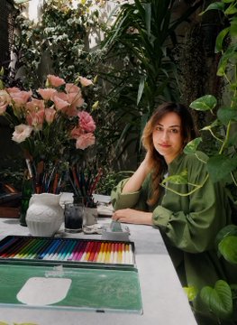 A woman in a green shirt sits beside a workspace featuring a tray of colorful watercolors, brushes, and a bouquet of pink flowers in a lush, plant-filled setting.