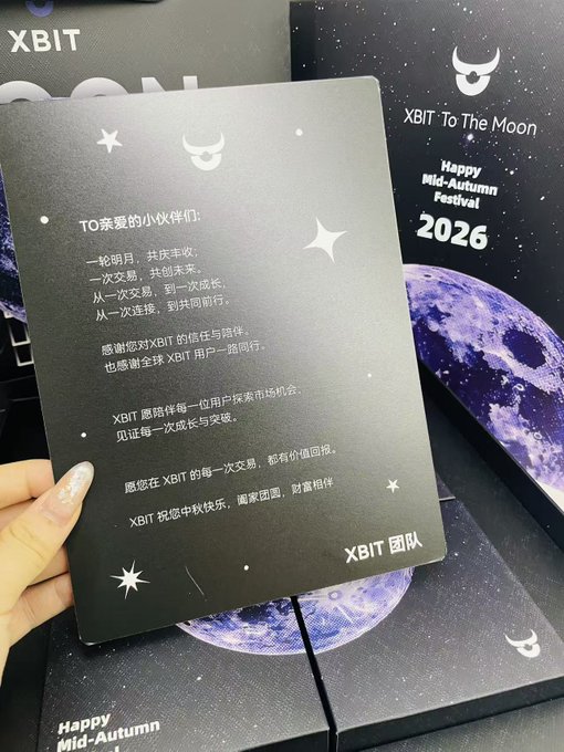 A hand holds a dark card with a Chinese message from the 'XBIT 团队' (XBIT Team) alongside boxes labeled 'XBIT To The Moon' and 'Happy Mid-Autumn Festival 2026'.