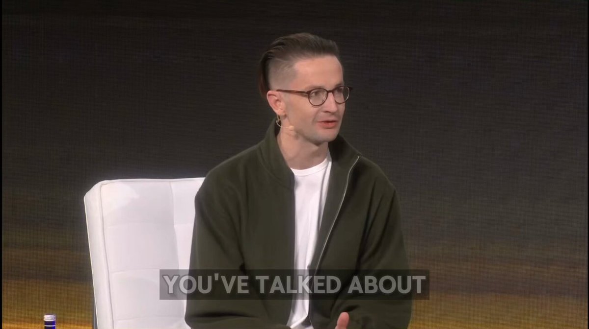 The image shows a man sitting down while talking about the global economy. The man has short hair and is wearing glasses. He appears to be engaged in conversation, possibly discussing the stock market or other financial topics.