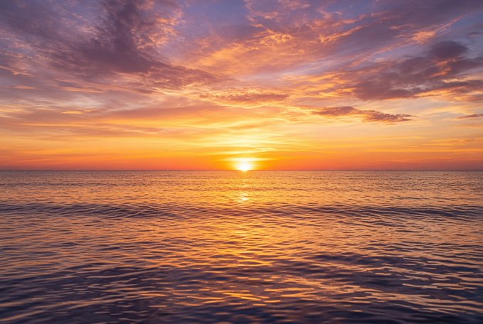 A scenic photograph of a vibrant orange and pink sunset over a calm ocean, with the sun's reflection stretching across the rippling water.