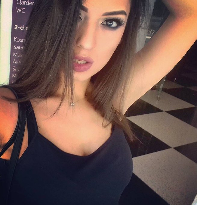 A selfie of a young woman with long brown hair, wearing a black tank top and a delicate necklace, posing in what appears to be a public restroom with a black and white checkered floor.