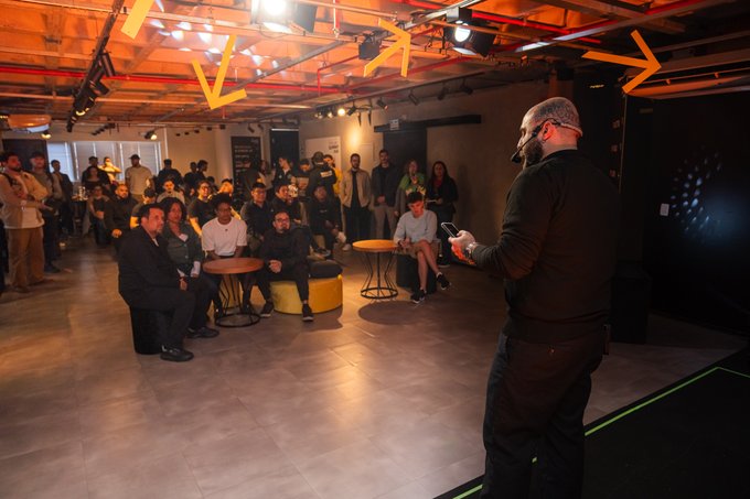 A presenter in dark clothing addresses a seated audience in a modern industrial-style venue with exposed ceiling pipes, orange arrow decorations, and warm ambient lighting.