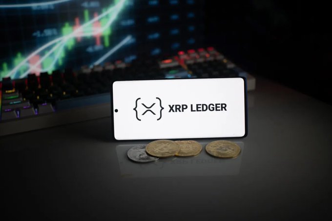 A smartphone displaying the 'XRP LEDGER' logo rests against a backdrop of financial trading charts and gold/silver coins on a dark surface.