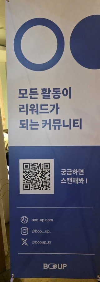 A blue and white promotional banner for 'BOOUP' (boo-up.com) with Korean text stating '모든 활동이 리워드가 되는 커뮤니티' (a community where all activities become rewards), featuring a QR code and social media handles.
