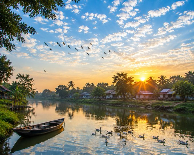 A serene landscape at sunrise showing a wooden boat on a calm river, ducks swimming, a flock of birds in the sky, and a village with palm trees in the distance.
