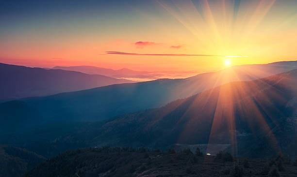 The image features a beautiful sunset over mountains with the sun's rays shining through them. The sky has hues of orange, yellow, and blue, creating a stunning backdrop for the scene. In the background, there are trees that add depth and texture to the landscape. The photo is taken from an elevated position, providing a bird's eye view of the breathtaking scenery below.