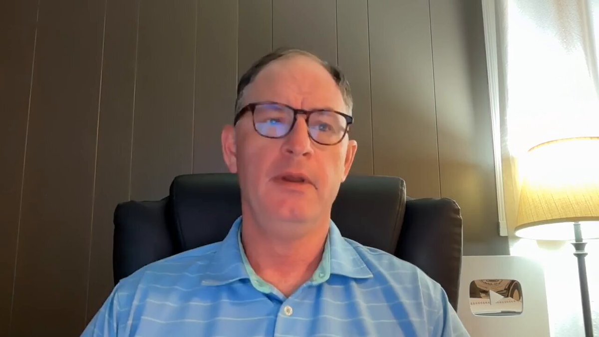 The image shows a man wearing glasses sitting in an office chair, looking at the camera with a serious expression. There are two lamps in the room - one near the left side of the frame and another towards the right side. The background features a wall with vertical wooden panels, which adds to the overall atmosphere of the scene.